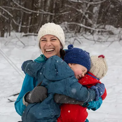 A woman in a blue coat has a beaming smile while hugging two children in red and blue snowsuits while playing outdoors in a wintry park.