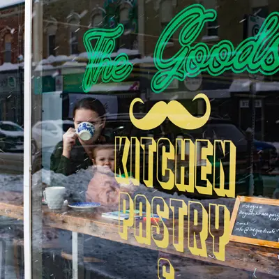 A photo of a woman holding a baby while enjoying a coffee, sitting by the front window of The Goods cafe.