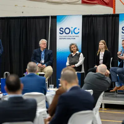 An audience listens to a panel of experts speaking about medical isotopes in front of a banner with the Southwestern Ontario Isotope Coalition logo.